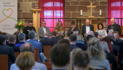 Mehrere Personen sitzen auf der Bühne mit Kerzen, einem Holzkreuz und einem Plakat bei der Fürbitte vor dem vollbesetzten Saal beim Zusammenschluss von Diakonie Werratal und Immanuel Diakonie Südthüringen im Kressehof im Meininger Ortsteil Walldorf 